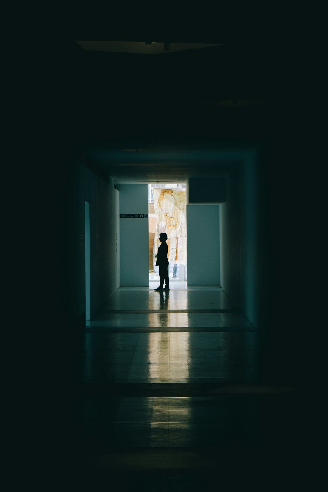A lone visitor silhouetted in a lit doorway at the end of a dark gallery corridor