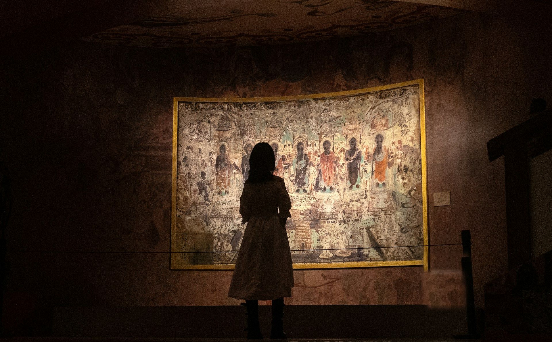 A visitor in silhouette standing before a large illuminated mural in a dark gallery