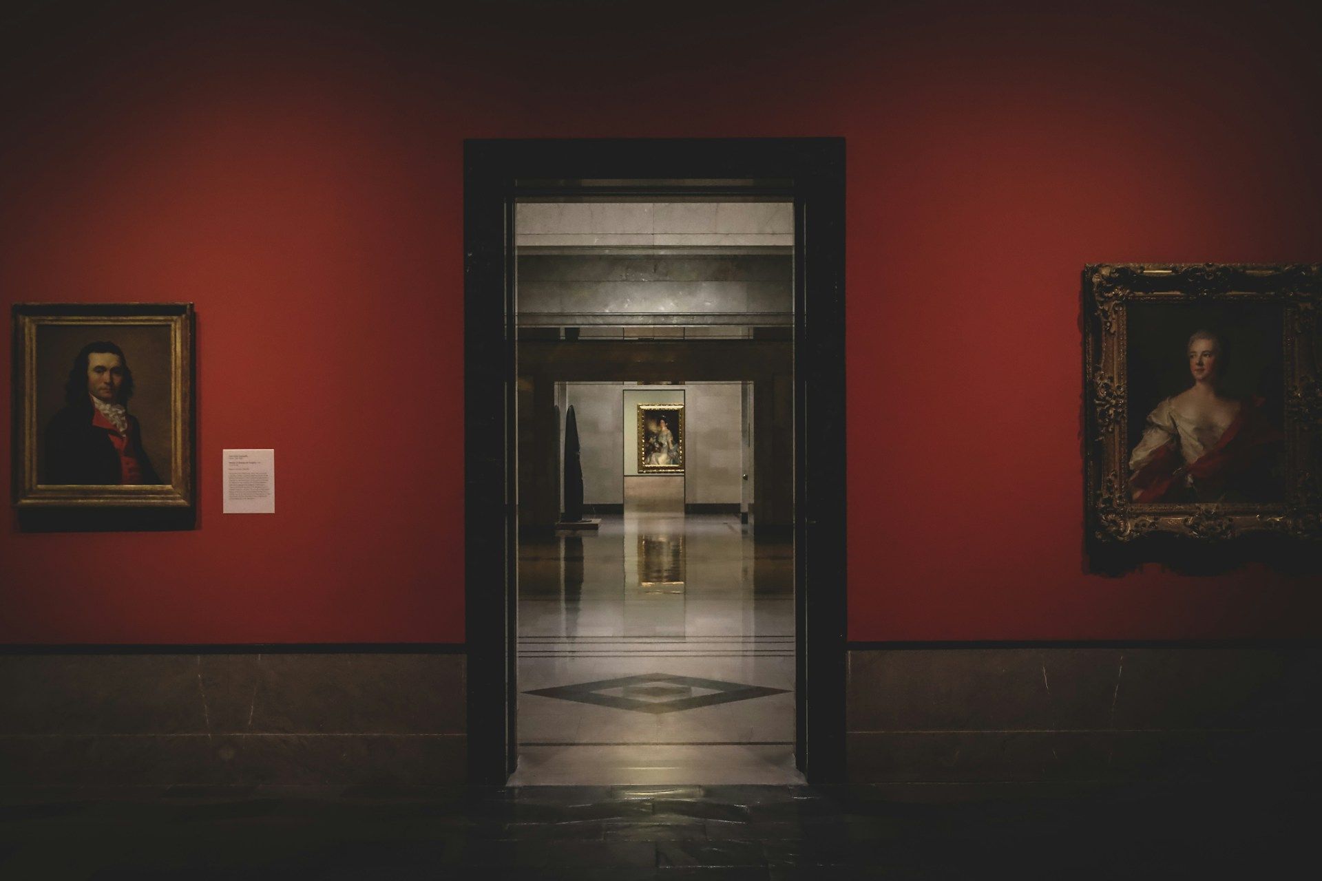 A doorway in a red gallery wall hung with portraits, opening onto a long lit corridor of galleries