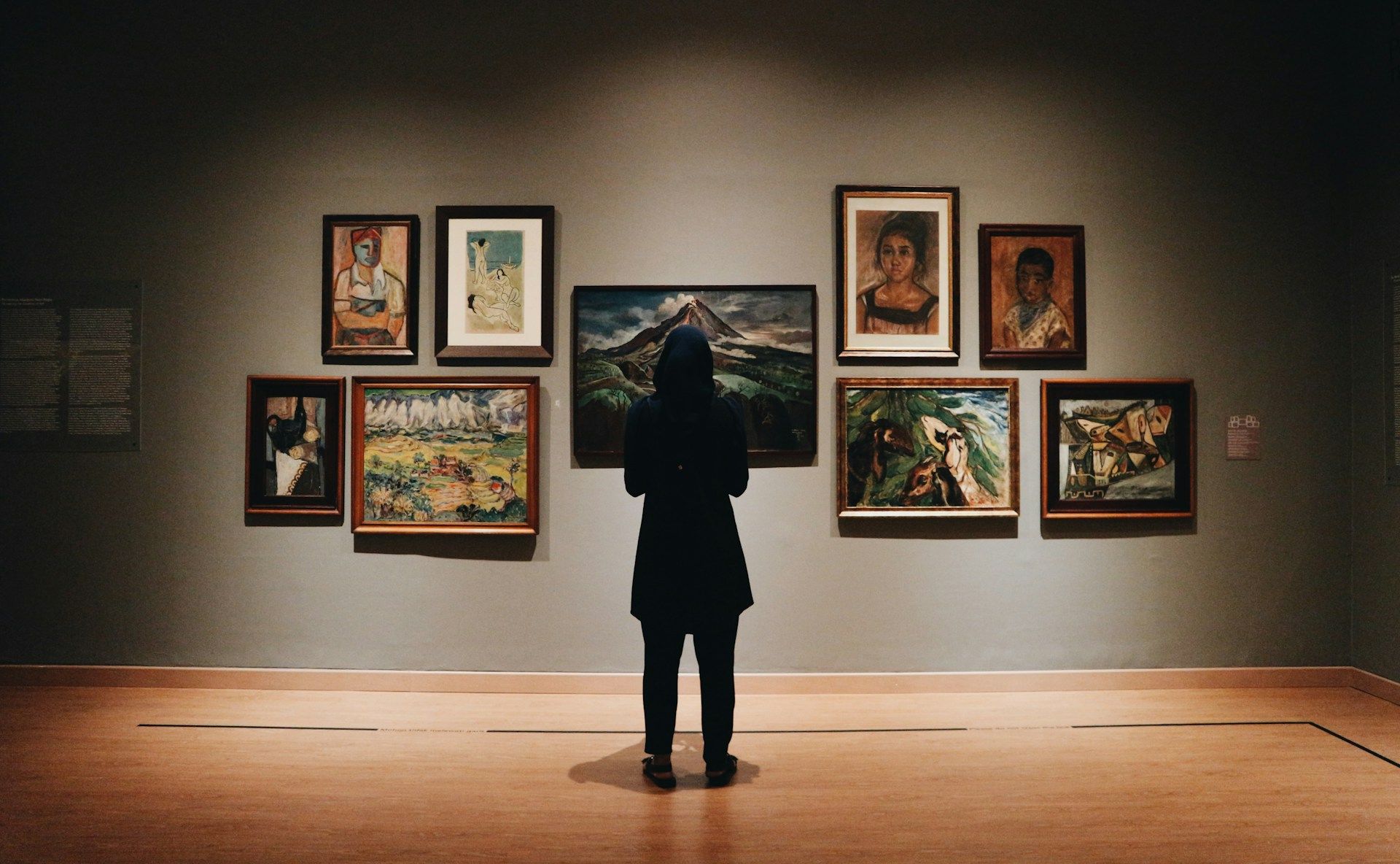 A visitor in a dark coat stands before a gallery wall hung with framed paintings