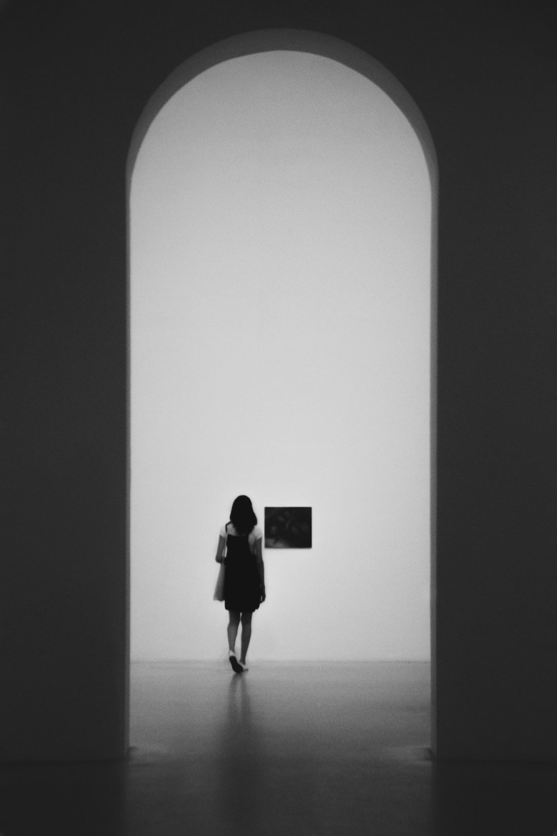 A lone visitor walks through a tall arched doorway toward a single painting on a pale wall