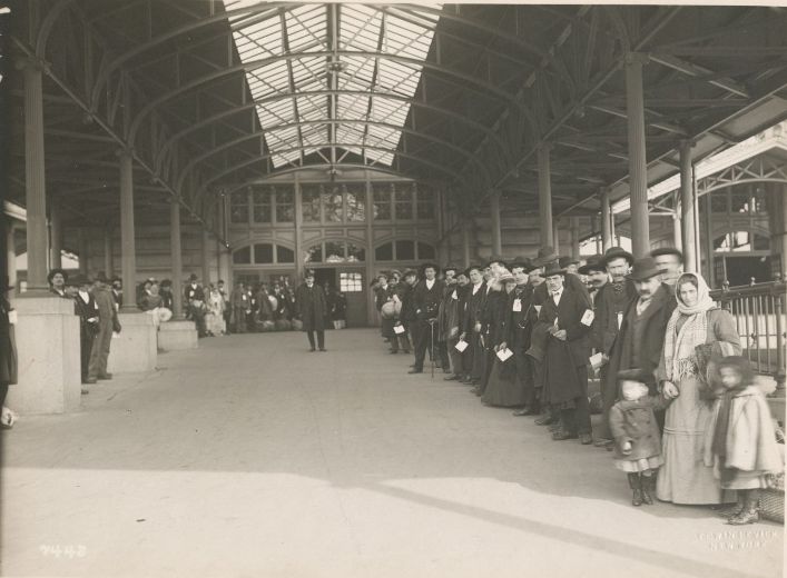 Immigrants, many holding papers, stand in a long line beneath a glass-roofed canopy