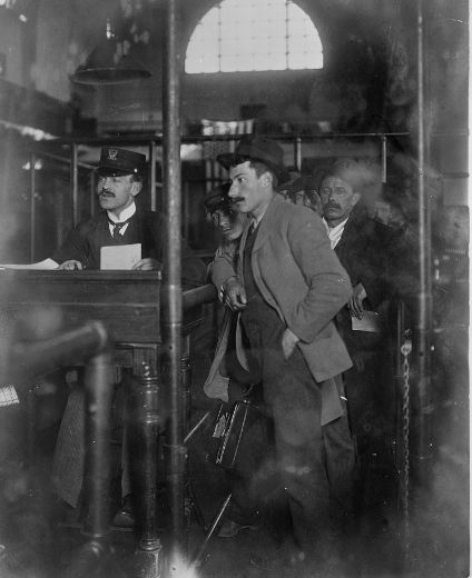 A man waits with others queued behind him at the registration desk at Ellis Island, where an official is seated