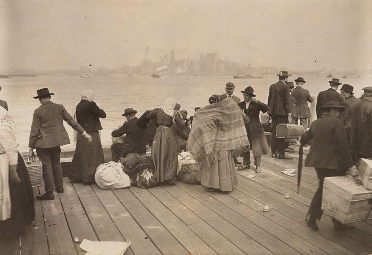 Men and women with bundles and suitcases wait on a dock, with New York City across the river